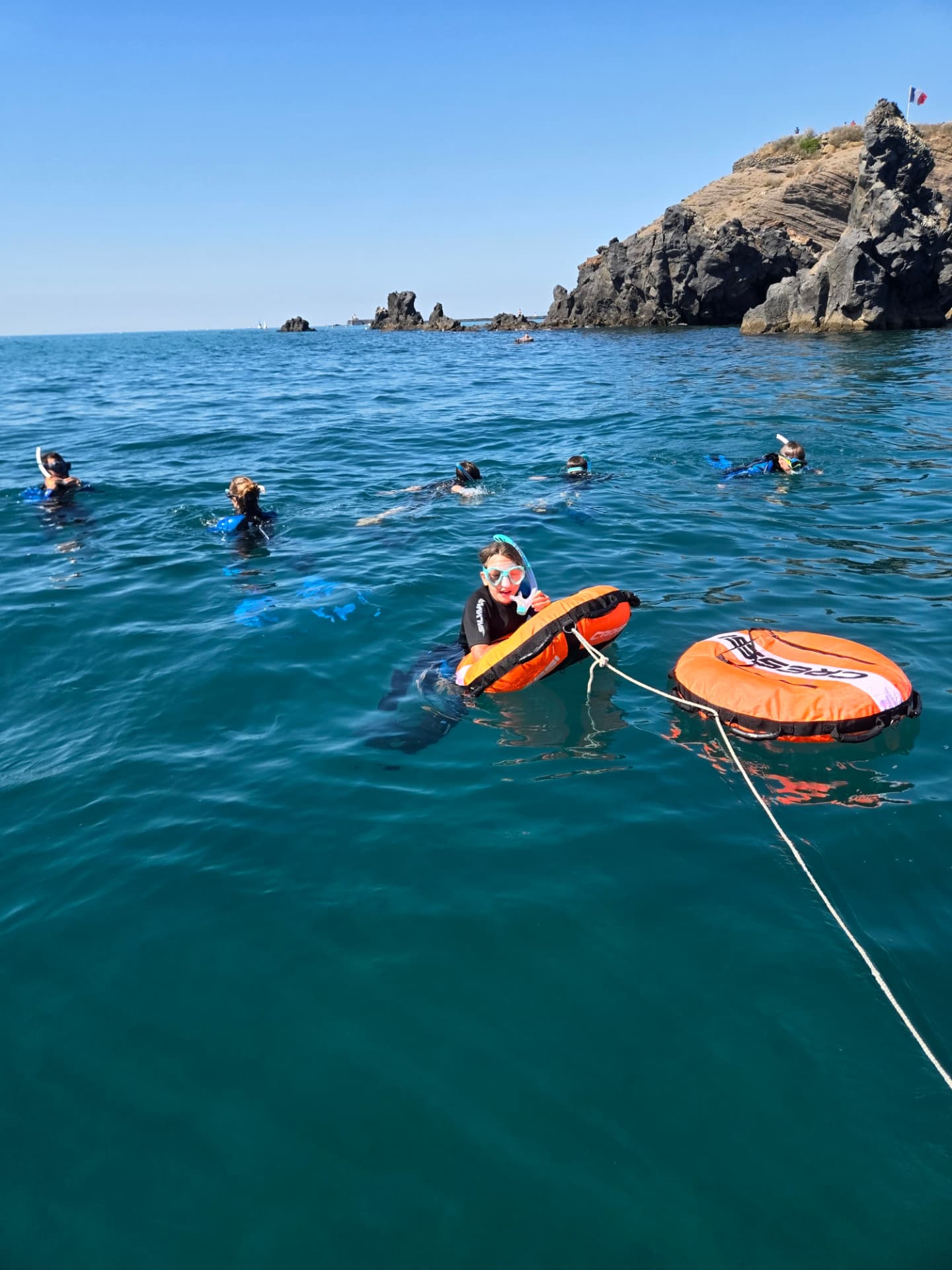Randonnée palmée : un groupe en masque et tuba, une bouée orange à la main, dans l’eau turquoise devant la côte rocheuse
